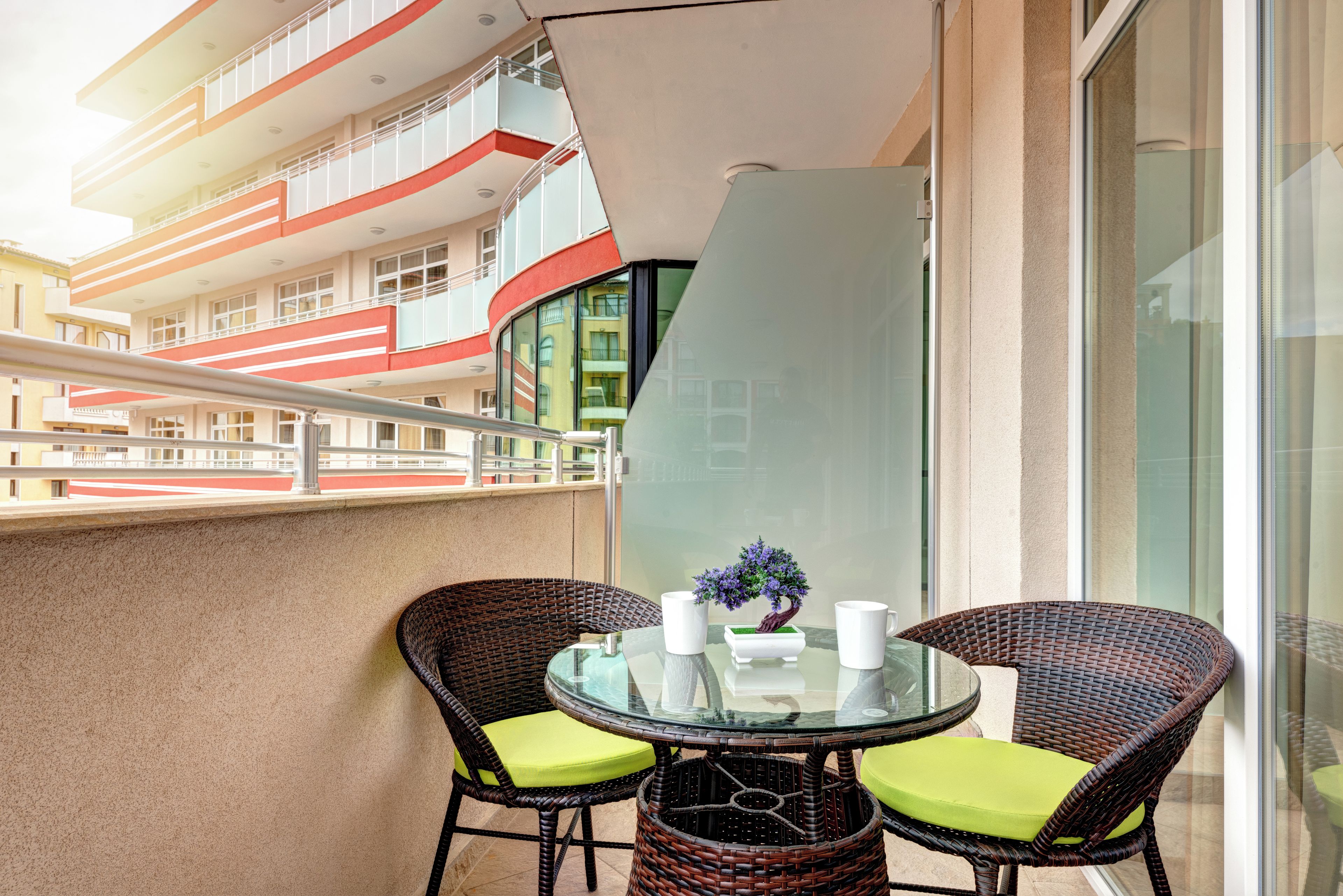 Un balcon doté de fauteuils en osier et d’une table en verre, offrant une vue sur un immeuble moderne également pourvu de balcons.