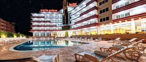 A hotel with a pool area, lounge chairs, and a night sky.