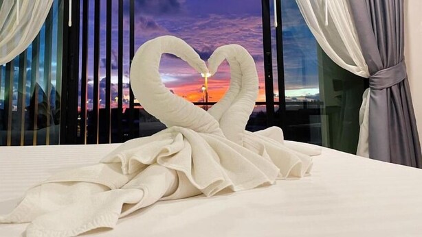 A bed with a towel arranged in a heart shape, a window with a sunset view, and curtains on the window. - Louis' Runway View Hotel (Mai Khao)