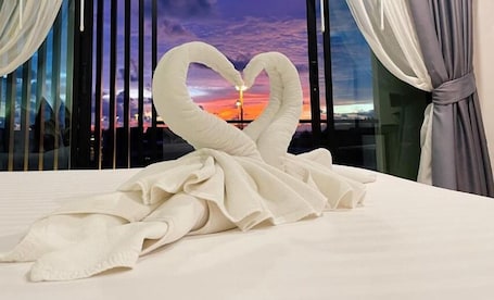 A bed with a towel arranged in a heart shape, a window with a sunset view, and curtains on the window.
