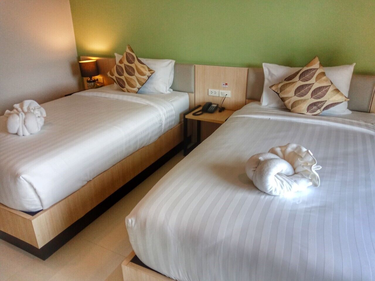 A hotel room with two beds, each with a pillow and a decorative item. There is a nightstand with a phone and a lamp between the beds.