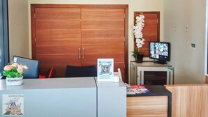 A hotel room with a wooden ceiling, a large wardrobe, a desk with a computer and a plant, a TV on a stand, and a QR code on the desk. - Louis' Runway View Hotel (Mai Khao)