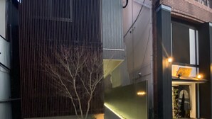A modern building with a metal facade, a small garden area with a tree, and a well-lit entrance. - Grand Base Nagasaki (Nagasaki)