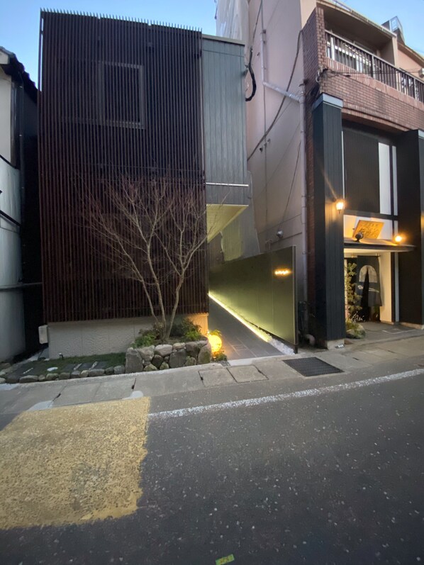 Un edificio moderno con fachada metálica, un pequeño jardín con un árbol y una entrada bien iluminada.