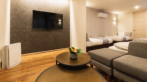 A modern hotel room with a flat-screen TV mounted on the wall, a round coffee table with a flower vase, a sofa, and a bed with two pillows. - Grand Base Nagasaki (Nagasaki)