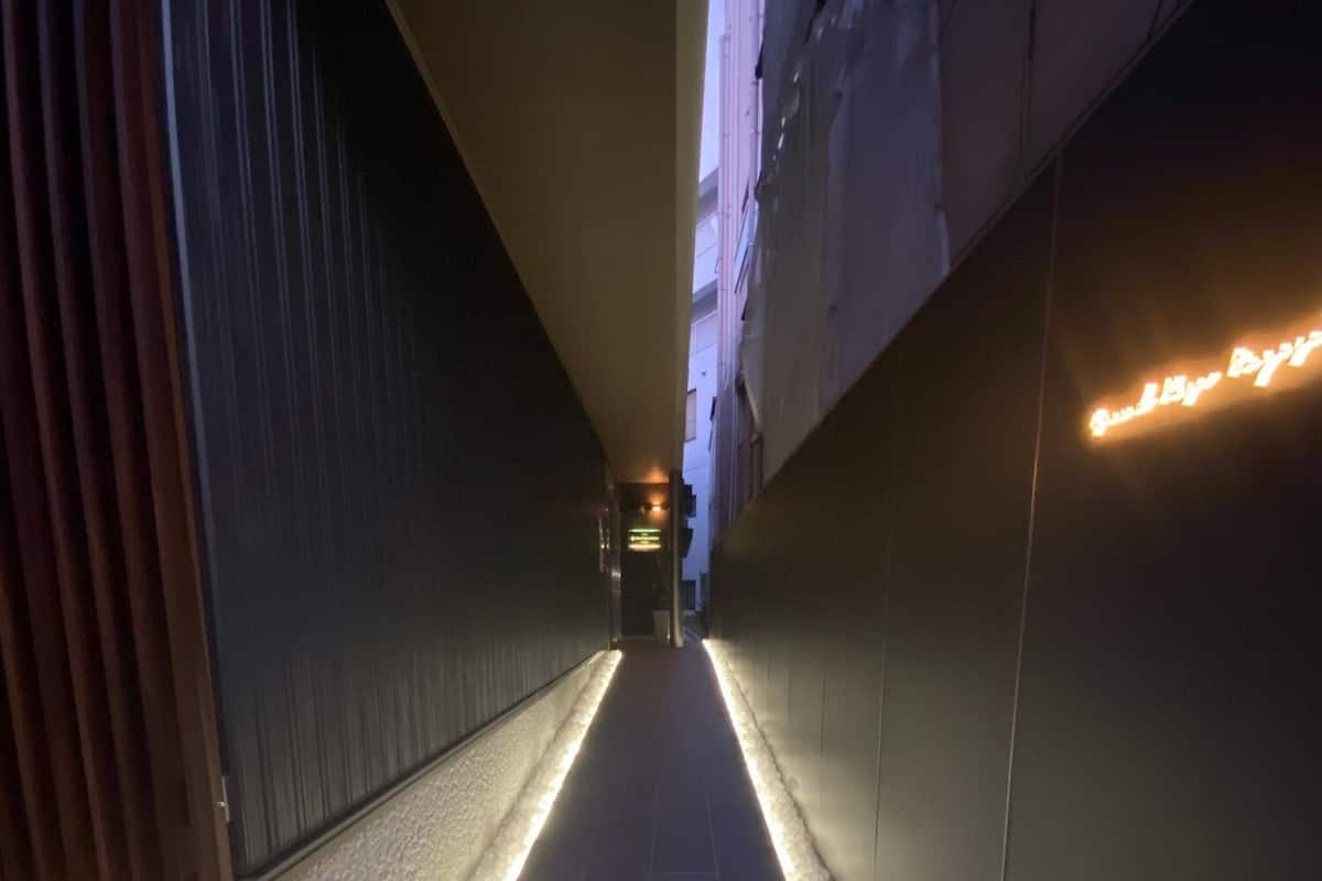 A narrow pathway with illuminated edges, a stone wall on one side, and a building with a neon sign on the other.