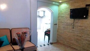 A living room with a sofa, a vase with flowers, a flat-screen TV mounted on a brick wall, and a doorway leading to a kitchen. - Nice apartment by the lake 4579 (Bin El Ouidane)