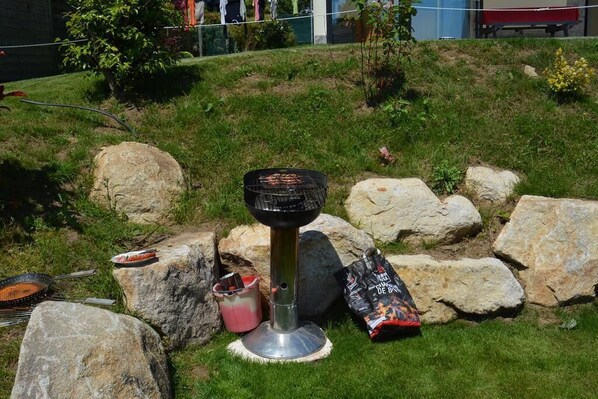 A portable grill setup on a grassy area with rocks, a bag of charcoal, and a grill pan.