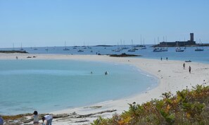 A beach with clear water, boats, and a distant building.