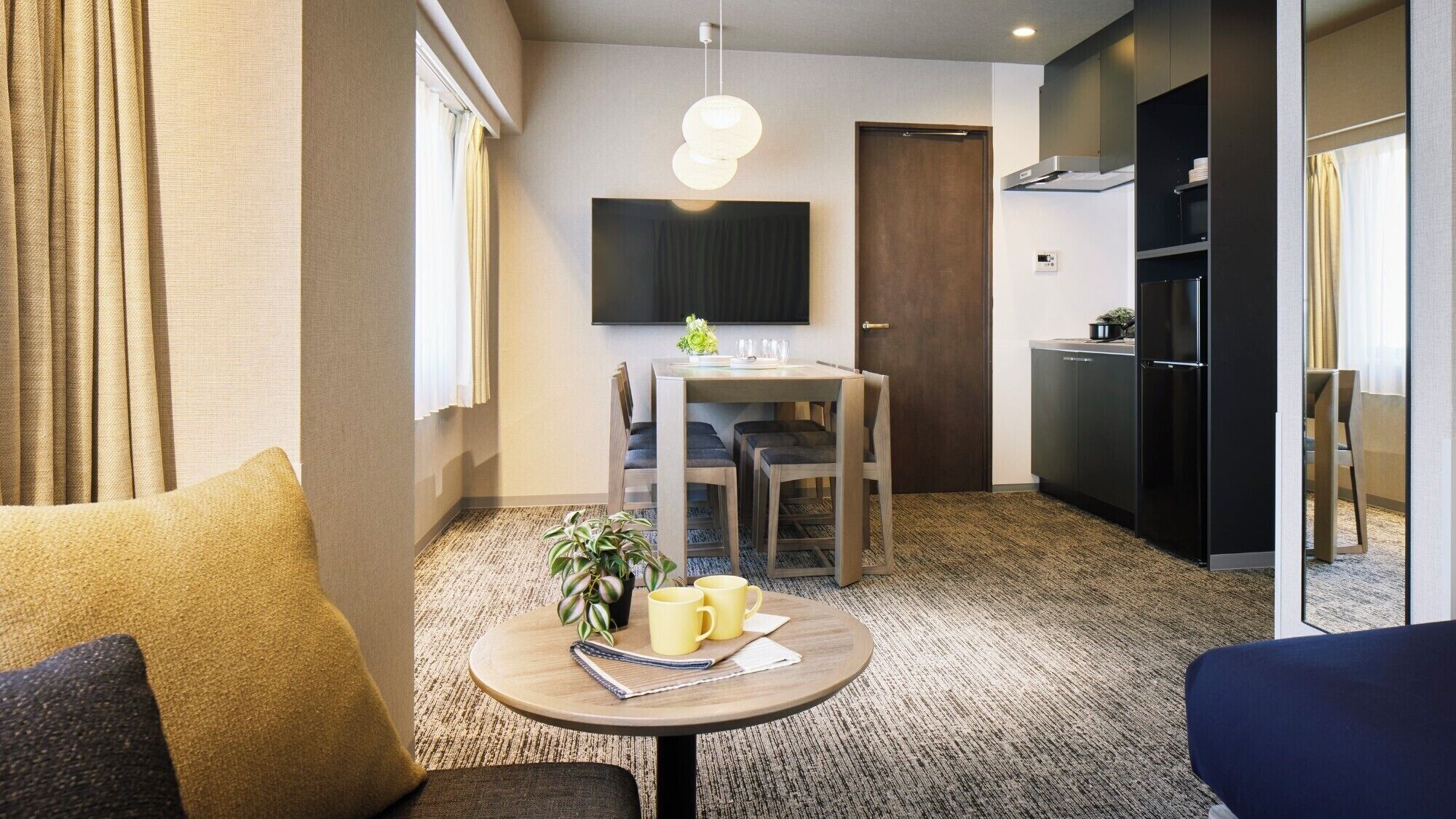 Una camera d'albergo moderna con zona pranzo, televisione a schermo piatto, angolo cottura con frigorifero e una grande finestra con tende.