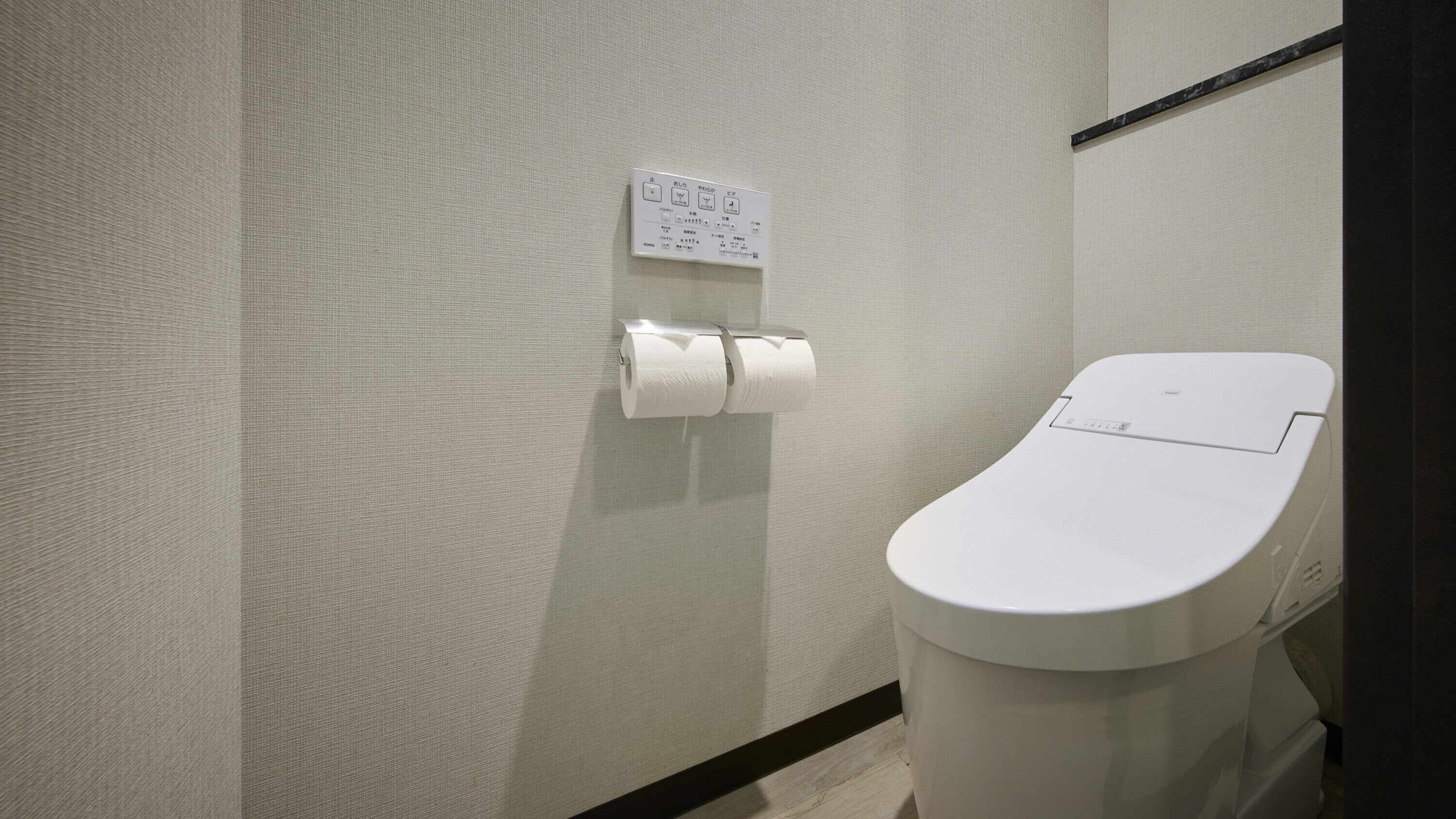Et toilet med hvidt sæde og låg, en toiletpapirholder med to ruller, og et skilt med japanske tegn hængt på væggen.