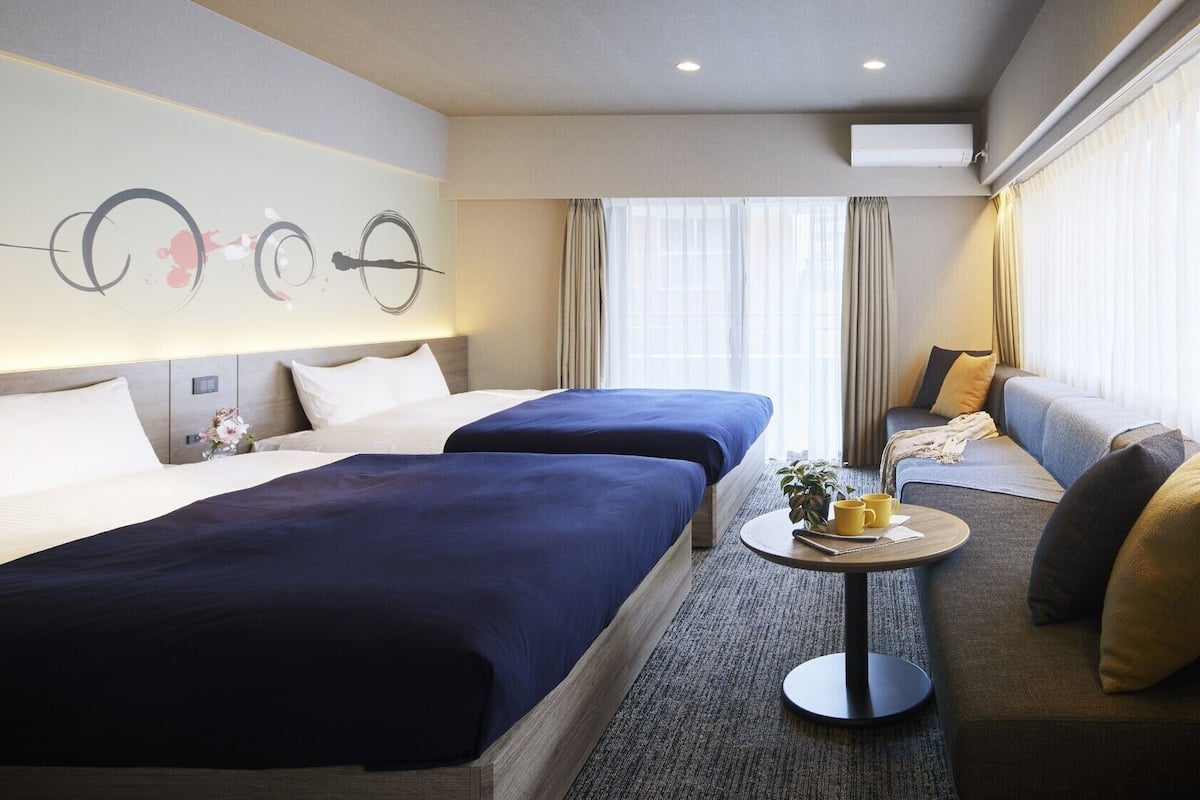 A hotel room with two beds, a sofa, a small table with a plant and a mug, and a large window with curtains.