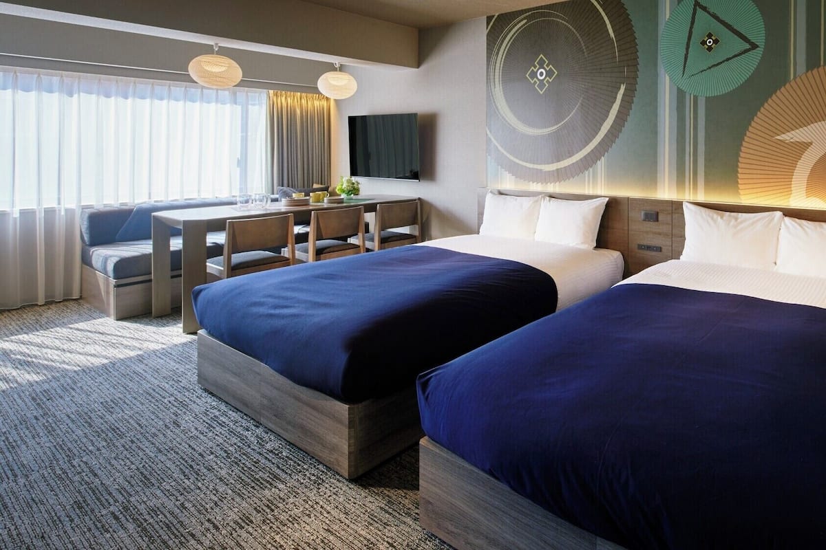 Une chambre d’hôtel avec deux lits, un bureau avec des chaises, un téléviseur à écran plat et des œuvres d’art décoratives au mur.