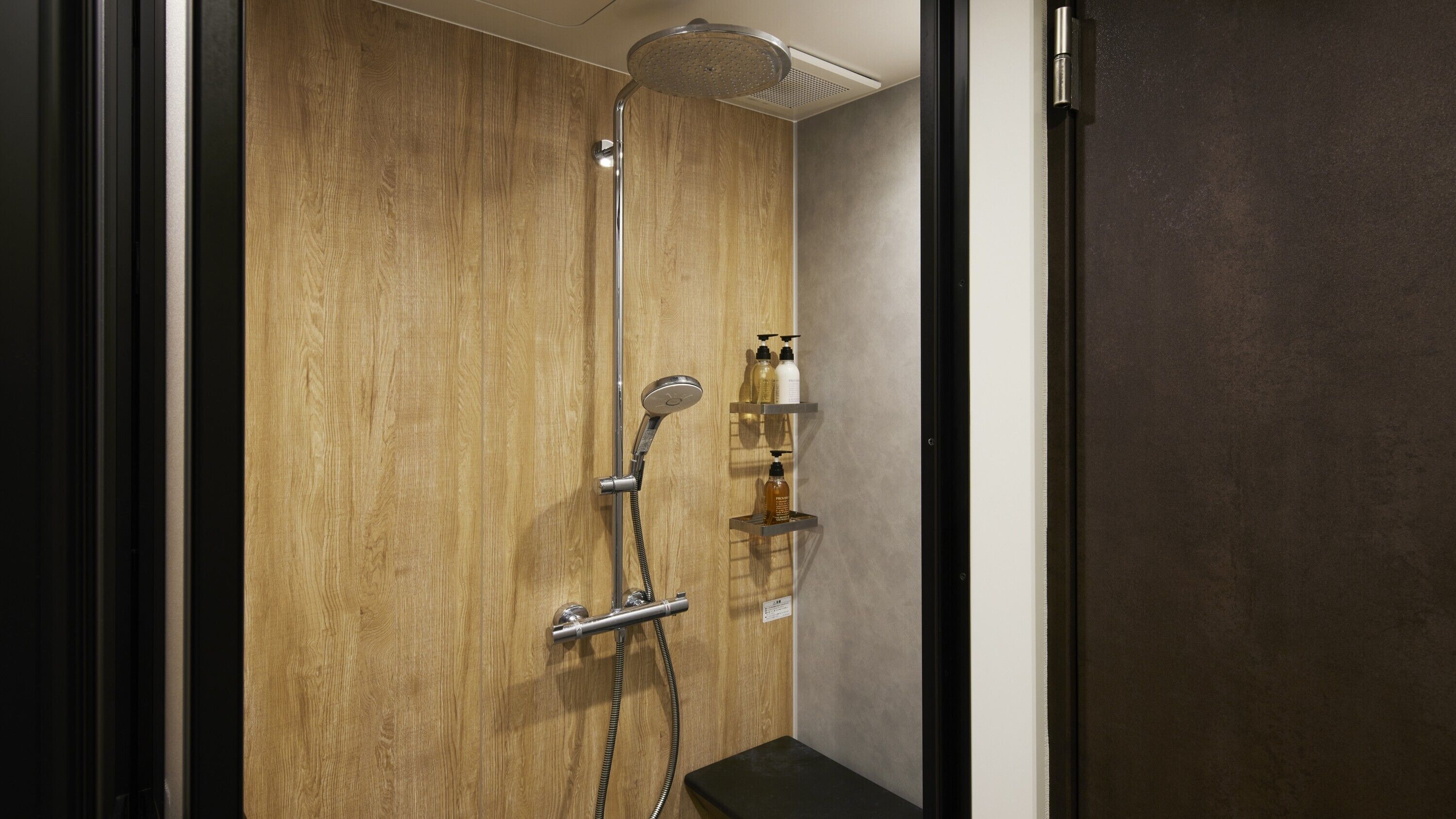 A shower with a wooden panel wall, a handheld showerhead, and a wall-mounted faucet. There are shelves with toiletries.