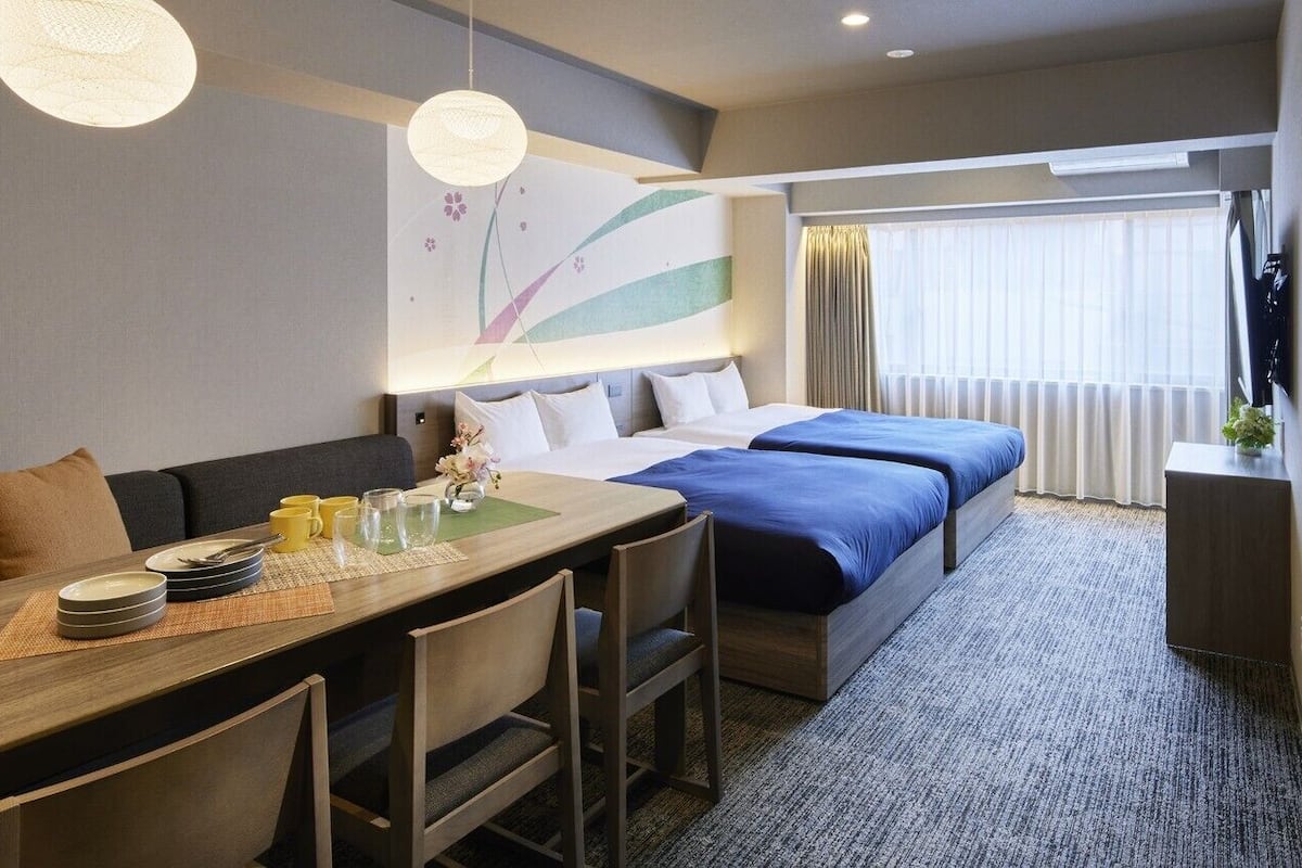 A hotel room with a dining table, chairs, a sofa, a bed with blue bedding, a large window with curtains, and a wall-mounted artwork.