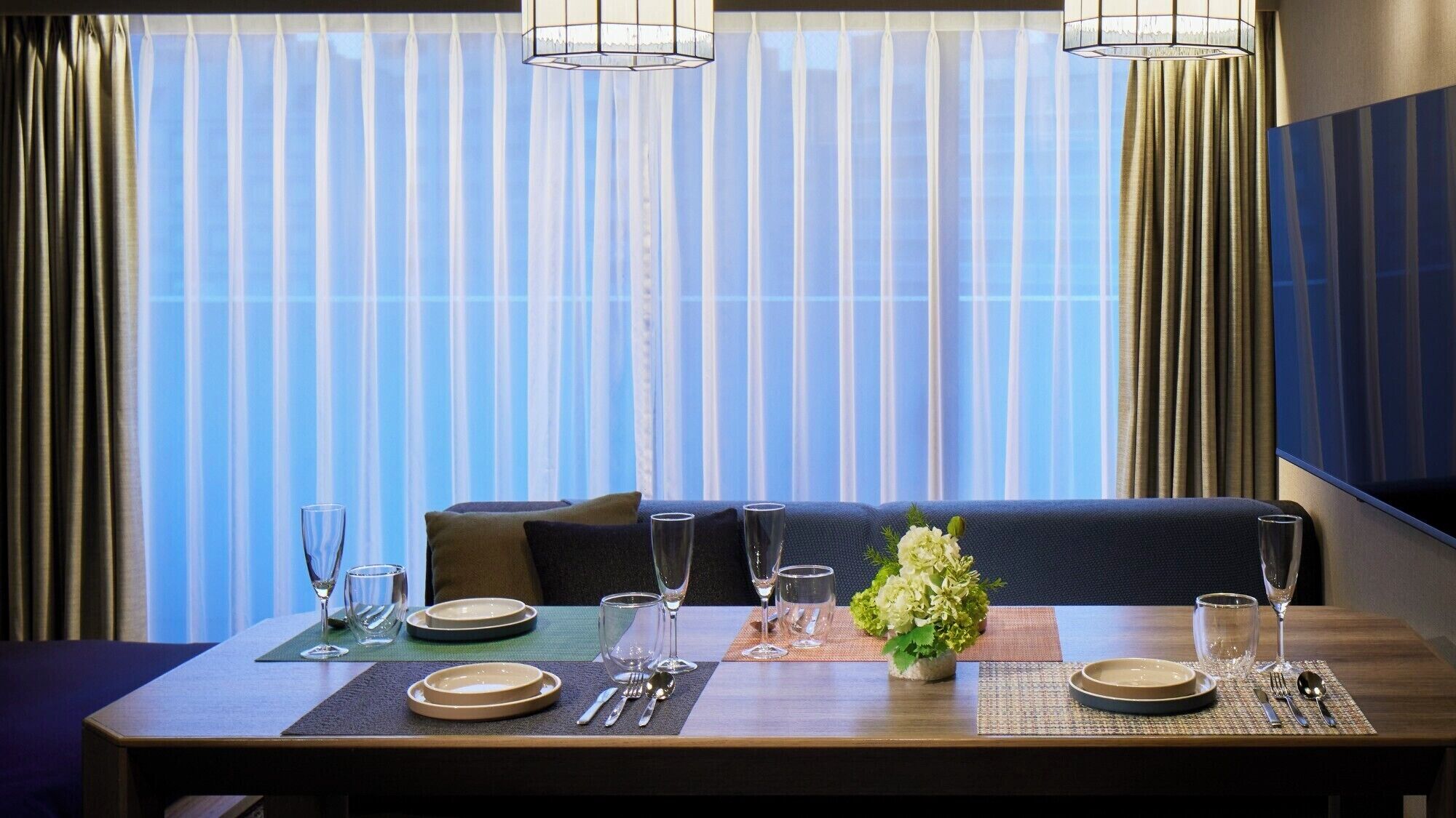 Un comedor con una mesa dispuesta para cuatro, incluyendo platos, cubiertos y vasos. Hay una planta en maceta sobre la mesa, y la habitación tiene un ventanal con cortinas.