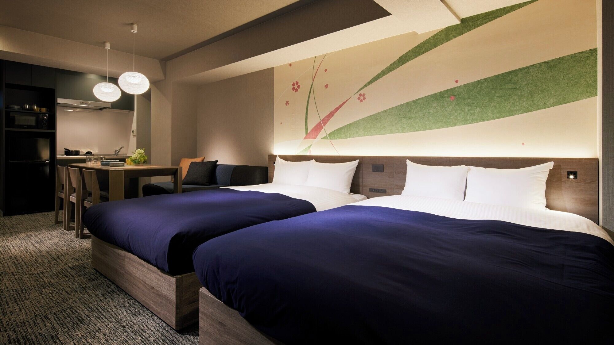 A hotel room with two beds, a desk, a chair, a small kitchenette, and a decorative wall.