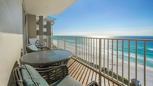 A balcony with a glass table and wicker chairs, overlooking a beach with clear turquoise waters.