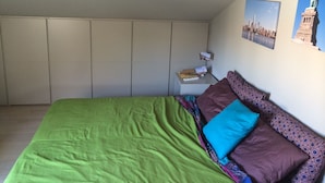 A bedroom with a green bedspread, a brown and blue pillow, a small bedside table with a remote control, and a wall with two framed pictures. - Rinidia Siena Celso Cittadini (Siena)