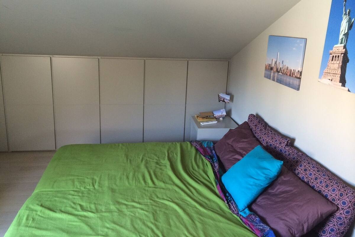 A bedroom with a green bedspread, a brown and blue pillow, a small bedside table with a remote control, and a wall with two framed pictures.