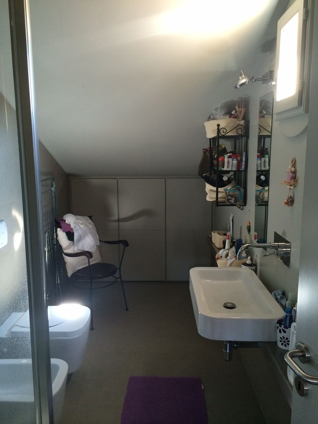 A compact bathroom with a white sink, a glass shower enclosure, a chair, and a small shelf with toiletries.