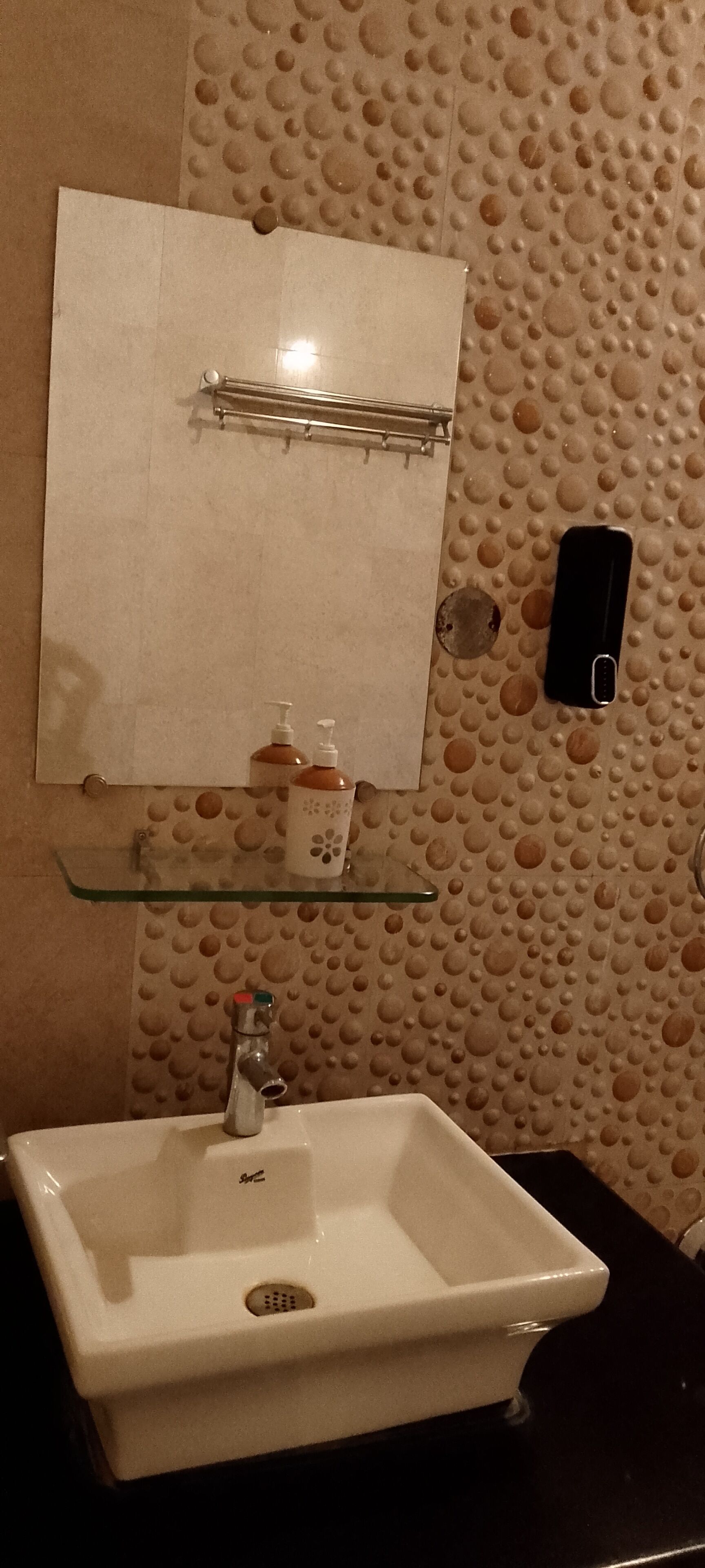 Un baño con un lavamanos blanco, un espejo y un dispensador de jabón sobre un estante de vidrio.