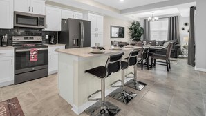 A modern kitchen with a central island, bar stools, a dining area with a sofa, and a living room with a TV.