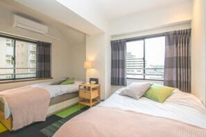Habitación de hotel con dos camas, una mesita de noche con lámpara y vistas a edificios a través de las ventanas.
