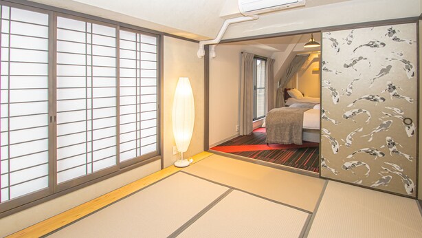 Una habitación de estilo japonés tradicional, con suelo de tatami, un panel corredizo shoji, una cama con detalles en rojo y una lámpara moderna.