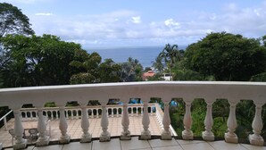 Varanda com grades brancas, com vista para o mar e vegetação exuberante.