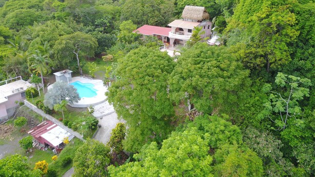 Uma casa cercada por floresta densa, com uma piscina visível e uma construção de telhado vermelho.