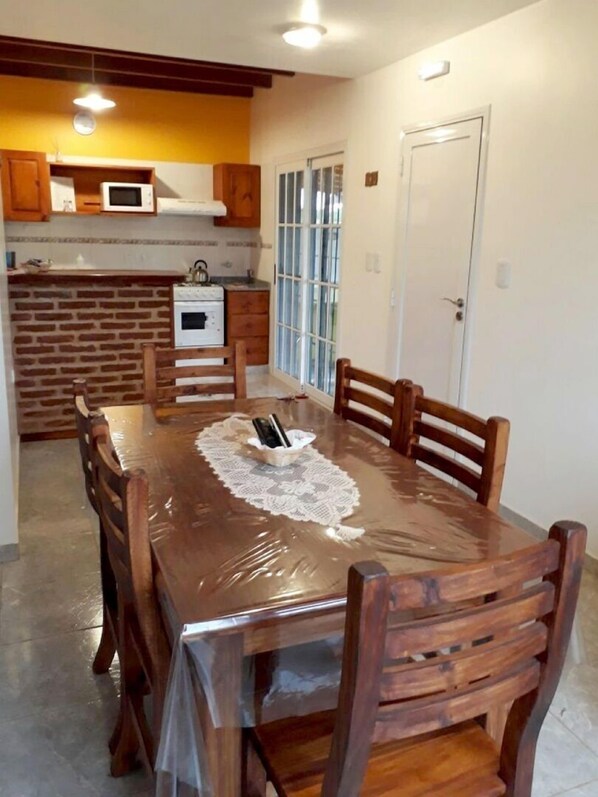 A dining area with a large wooden table covered with a plastic tablecloth, surrounded by wooden chairs. A microwave and oven are visible in the adjacent kitchen area.