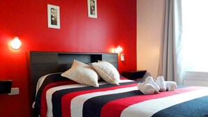 A hotel room with a red wall, a bed with a striped bedspread, two pillows, and two white towels on the bed.