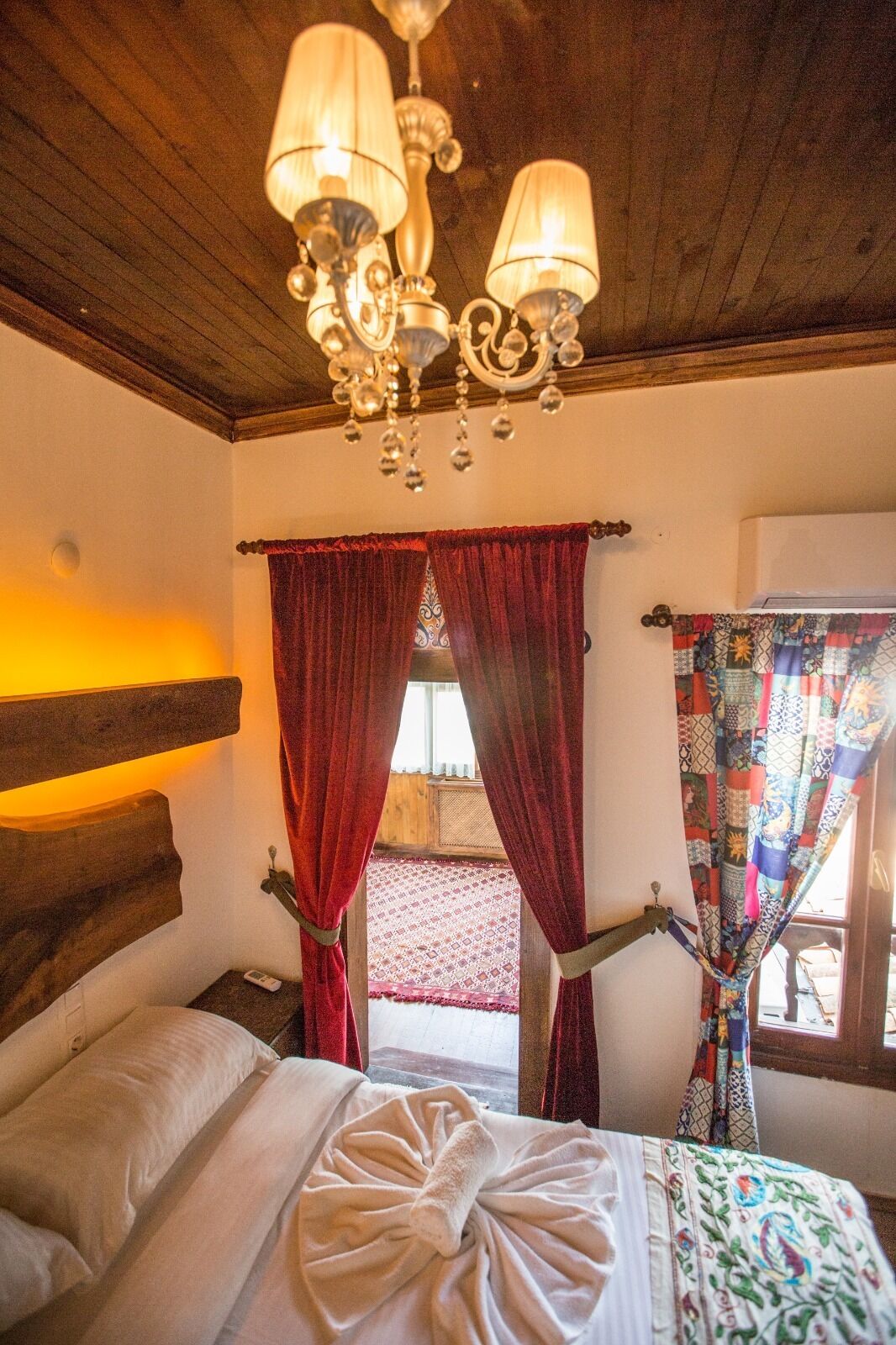 Un dormitorio con cabecera de madera, araña, cortinas rojas y cama con toalla blanca.