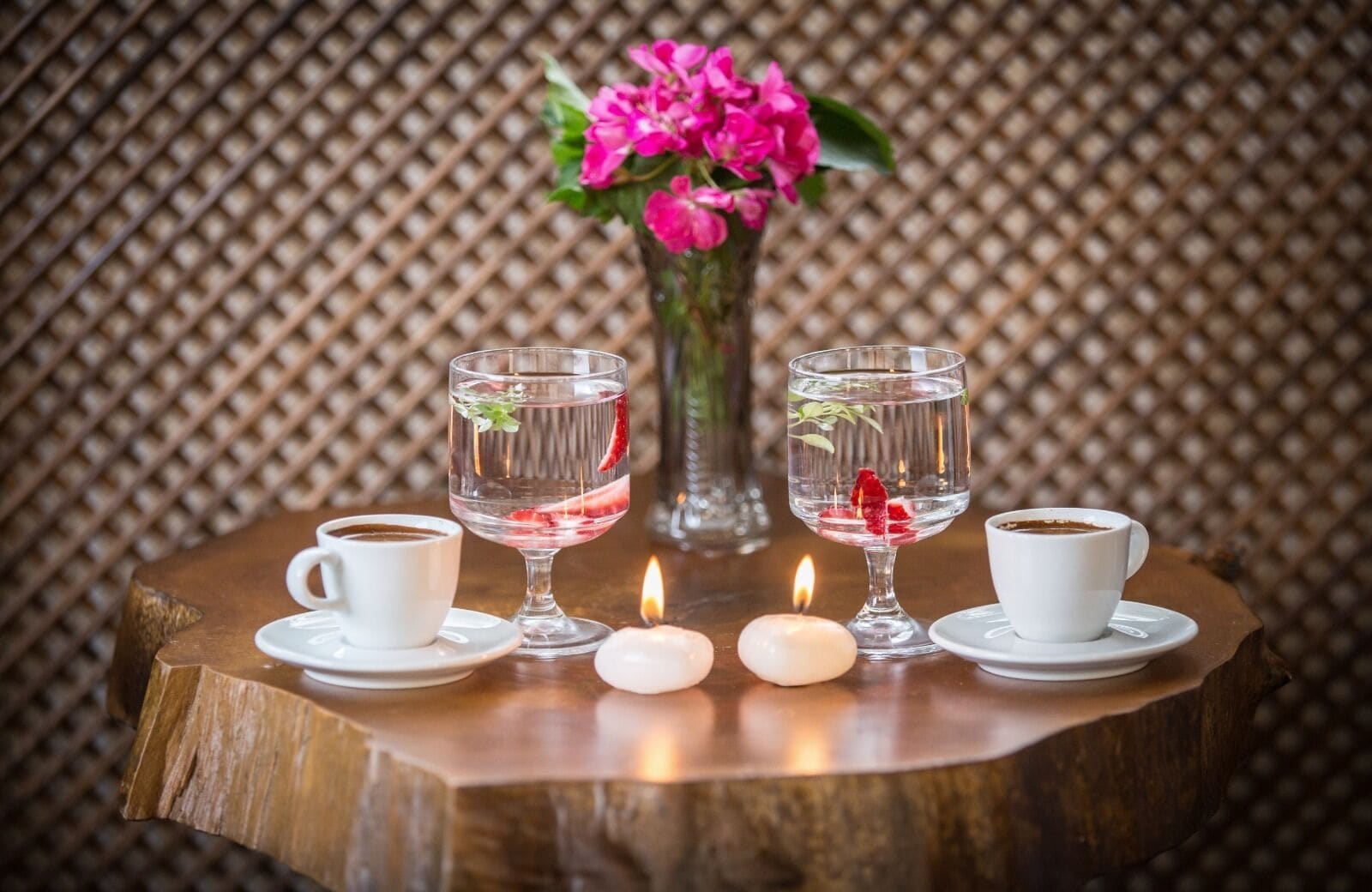 A table with two white cups of coffee, two clear glasses with water and strawberries, a vase with pink flowers, and two lit candles.