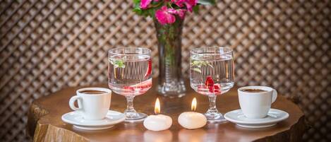 A table with two white cups of coffee, two clear glasses with water and strawberries, a vase with pink flowers, and two lit candles.