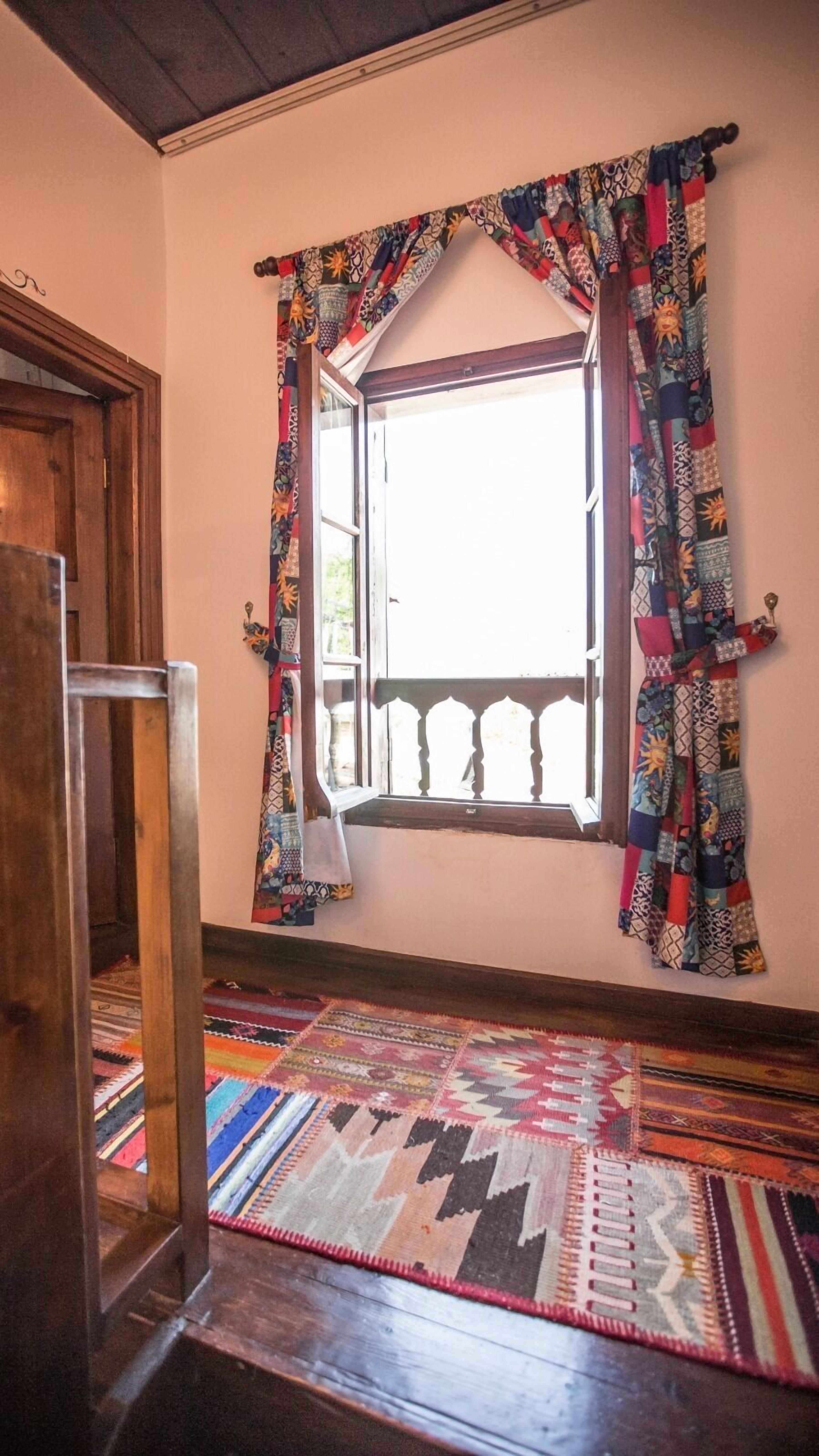 A room with a wooden floor, a window with patterned curtains, and a view of the outdoors.