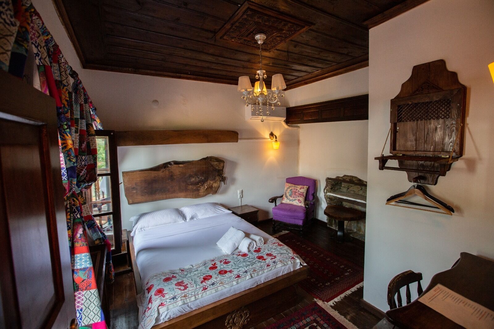 Un dormitorio con un cabecero de madera grande, una lámpara de araña colgante, un sillón morado y un perchero de madera.