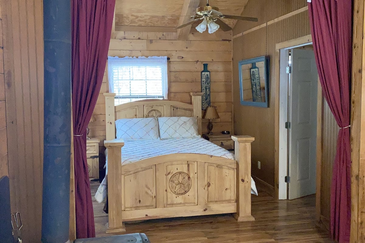A wooden cabin bedroom with a bed, curtains, a ceiling fan, and a rustic aesthetic.