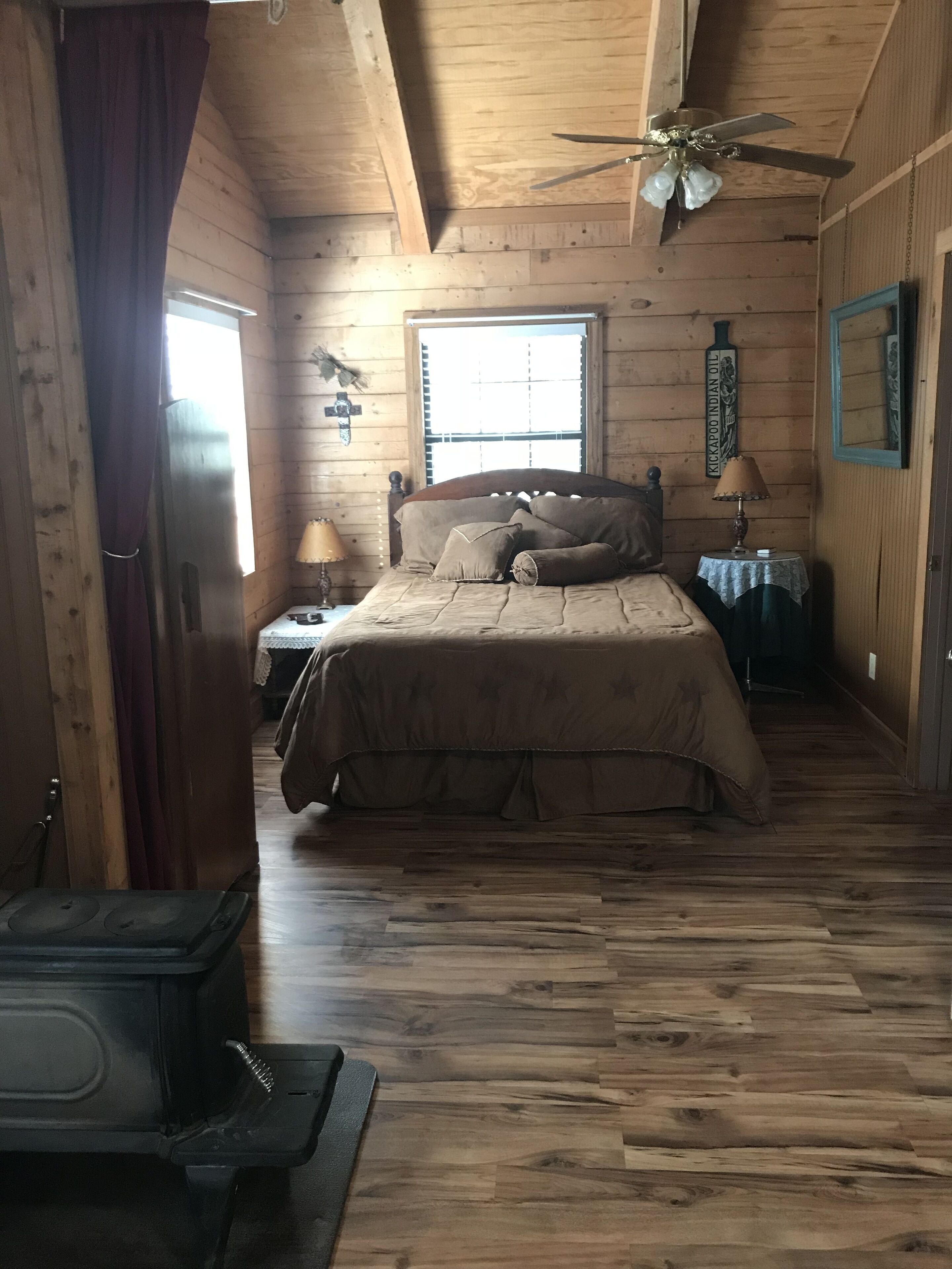 Un dormitorio de cabaña de madera con una cama, un mesita de noche, una lámpara, un ventilador de techo y una ventana.