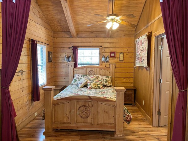 A wooden cabin bedroom with a bed, curtains, a ceiling fan, and a window.
