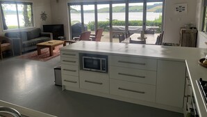 A modern kitchen with a central island, large windows offering a view of the outdoors, and a dining area with chairs and a table. - Laze by the Lake - Holiday Home - Stunning Views of Lake Rotoiti  (Tikitere)