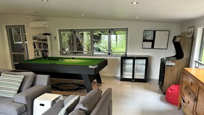 A modern living room with a pool table, grey sofas, a red bean bag, a wooden cabinet, and a large window with a view of greenery. - Laze by the Lake - Holiday Home - Stunning Views of Lake Rotoiti  (Tikitere)