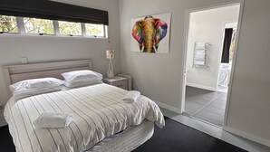 A modern bedroom with a large bed, a nightstand, a wall-mounted artwork, and a view of a bathroom through an open door. - Laze by the Lake - Holiday Home - Stunning Views of Lake Rotoiti  (Tikitere)