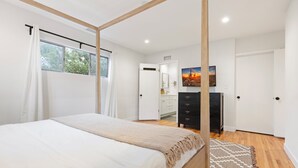 Une chambre à coucher avec un lit à baldaquin, un plancher en bois, un tapis à motifs, une commode, un tableau et une fenêtre avec des rideaux.
