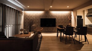 A modern living room with a flat-screen TV mounted on a textured wall, a dining area with a wooden table and chairs, and a kitchen area with a stove and microwave. - Crice Hotel Namba II (Osaka)