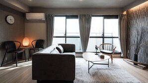 A modern living room with a sofa, two chairs, a coffee table, and a lamp. There is a clock on the wall and an air conditioning unit. - Crice Hotel Namba II (Osaka)