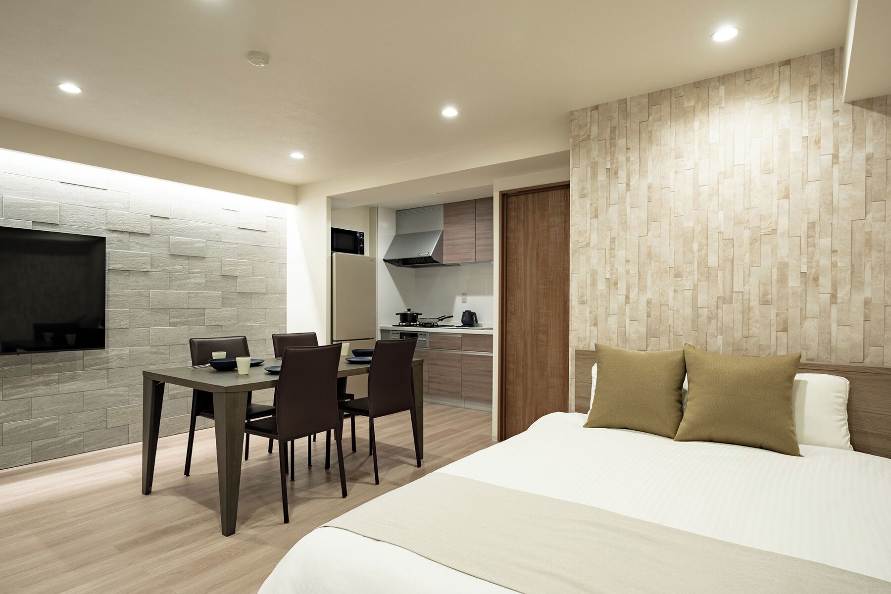 A modern bedroom with a large bed, a dining area with a table and chairs, and a kitchen in the background.