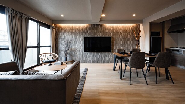 A modern living room with a sofa, a dining table with chairs, a flat-screen TV mounted on a textured wall, and a kitchen area with a stove and microwave. - Crice Hotel Namba II (Osaka)