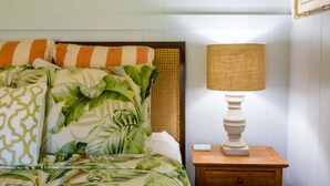 A bed with a tropical leaf pattern bedspread, a striped pillow, and a wooden nightstand with a lamp. - Waimea Beach Cottage by Destination Kauai - Charming 2BD, A/C, Walk to Ocean/Pier (Waimea)